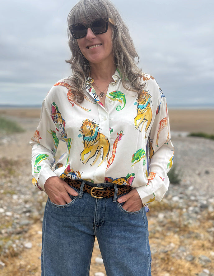 A woman wearing a white long-sleeve button-up shirt with a colorful safari animal print, paired with blue jeans and a leopard-print belt, standing outdoors on a rocky beach under an overcast sky.