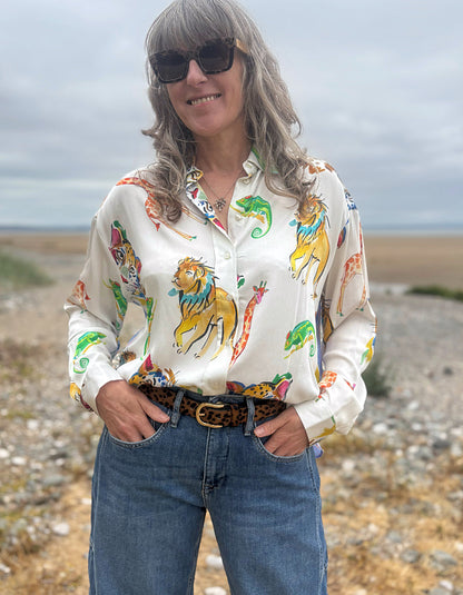 A woman wearing a white long-sleeve button-up shirt with a colorful safari animal print, paired with blue jeans and a leopard-print belt, standing outdoors on a rocky beach under an overcast sky.