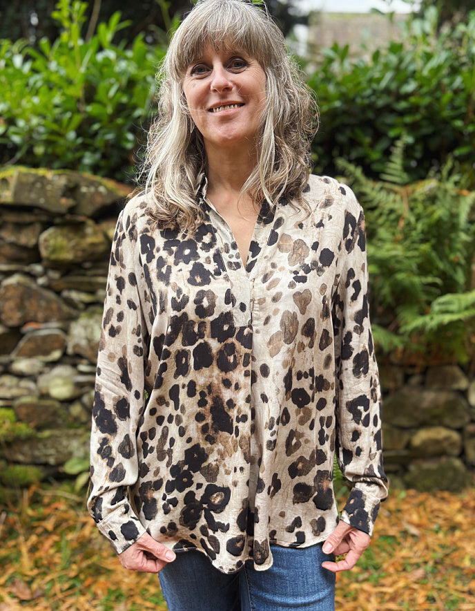 Woman wearing a leopard print shirt outdoors with greenery and a stone wall in the background