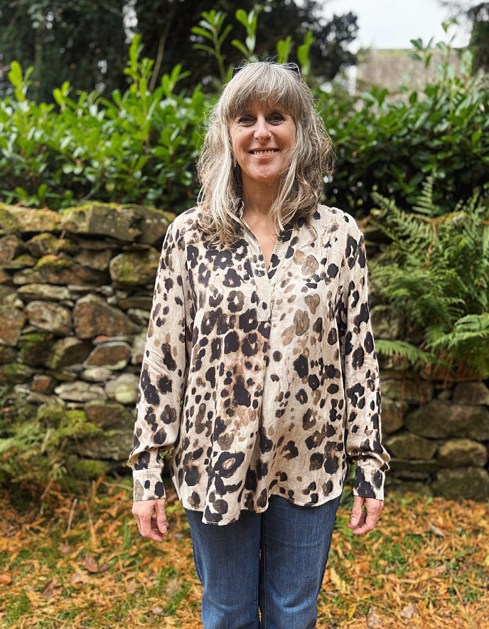 Woman wearing a leopard print blouse standing in a garden with a stone wall and greenery.