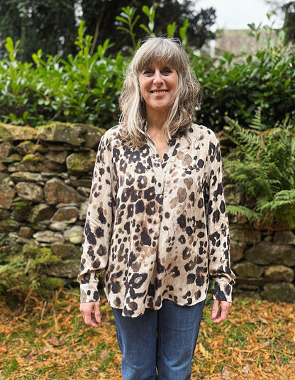 Woman wearing a leopard print blouse standing in a garden with a stone wall and greenery.