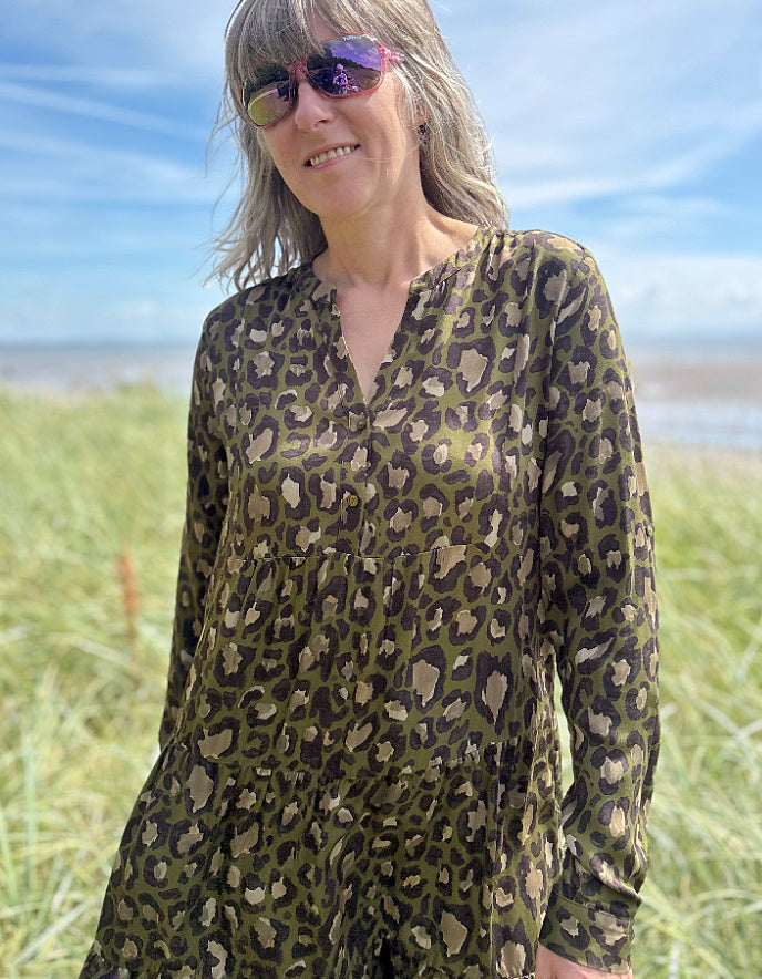 Person wearing a leopard print dress standing on a grassy field with a beach and blue sky in the background