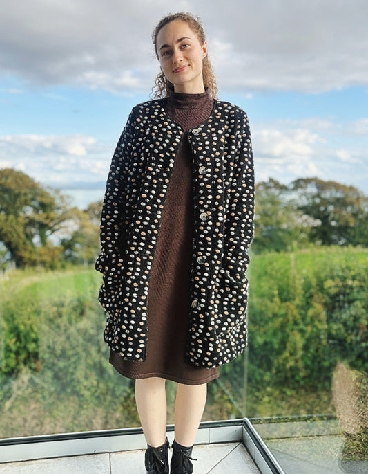 Woman wearing a black and white polka dot coat with a brown dress, standing outdoors.