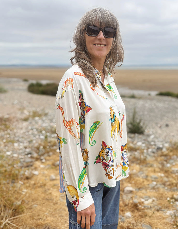 A woman wearing a long-sleeve button-up shirt with a colorful animal print, paired with blue jeans and sunglasses, standing outdoors in a rocky, dry landscape.
