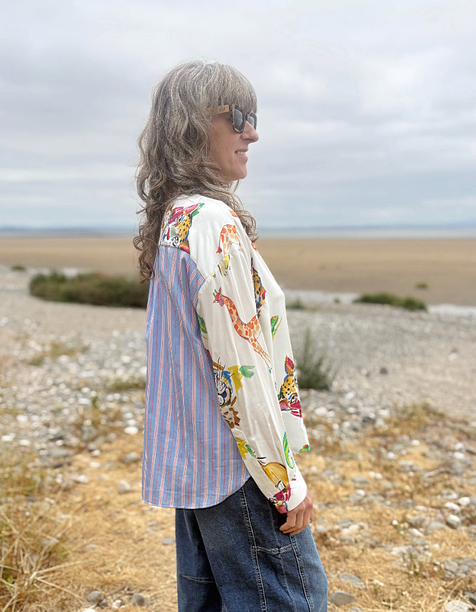 A woman with long gray hair and sunglasses stands in profile on a rocky beach, wearing a mixed-print shirt with blue and white vertical stripes on one side and a cream multicolor animal print on the other, paired with dark blue jeans.