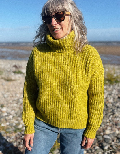 A woman with gray hair and sunglasses wearing a chunky ribbed mustard yellow turtleneck sweater and blue jeans, standing on a rocky beach with water and sky in the background.