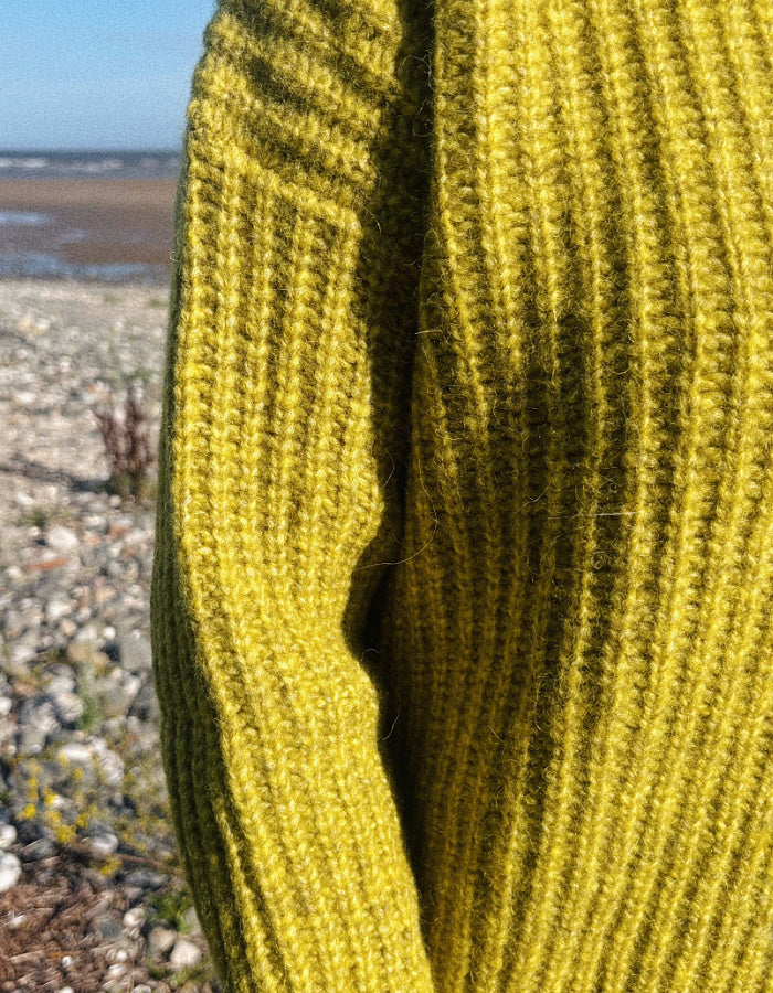 Close-up of a ribbed knit sweater in a mustard yellow color, shown outdoors with a blurred beach background.