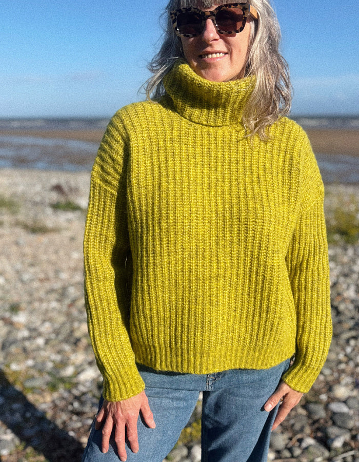 A woman wearing a chunky ribbed mustard yellow turtleneck sweater and blue jeans, standing outdoors on a rocky beach under a blue sky.