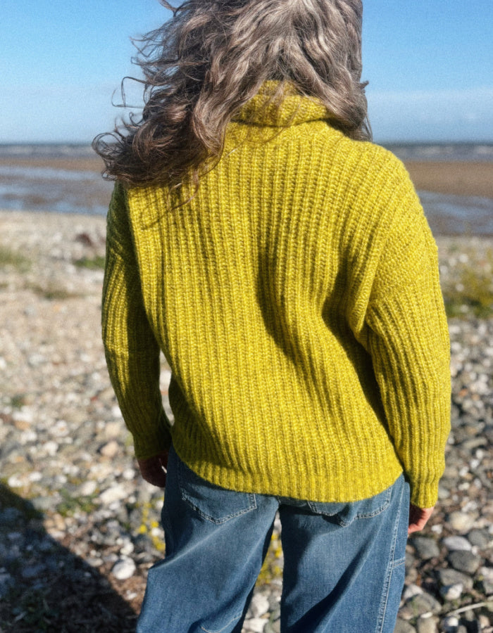 A woman viewed from behind wearing a bright yellow ribbed knit sweater and blue jeans, standing on a rocky beach with water and sky in the background.