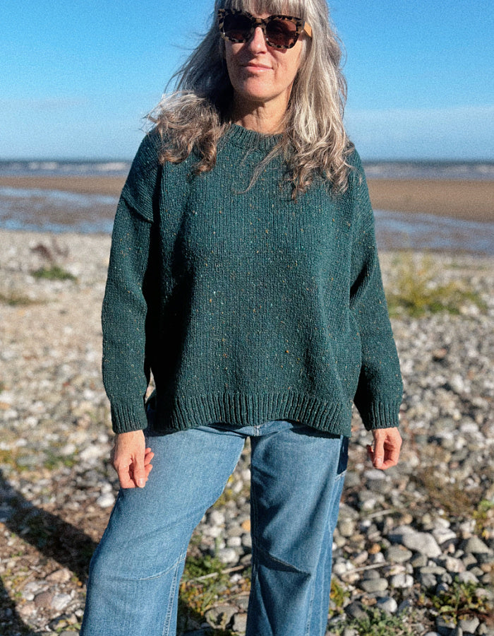 A woman wearing a dark green donegal tweed knit crewneck sweater and blue jeans stands on a rocky beach with the sea and blue sky in the background.