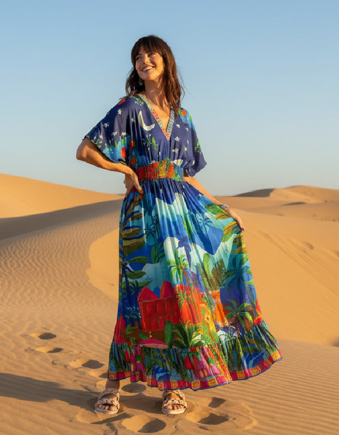 Woman in a colourful shirred waisted maxi dress with arabian nights print standing on sand dunes with a clear blue sky.