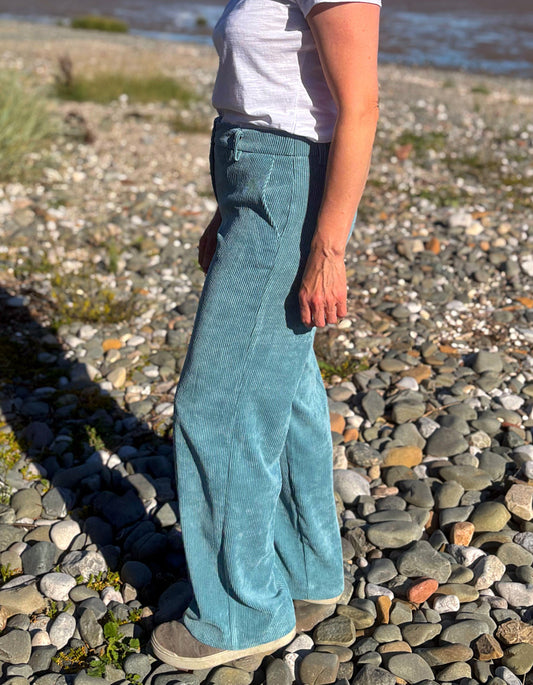 A person standing on a pebble beach wearing wide-leg teal corduroy pants, a white t-shirt, and tan shoes.