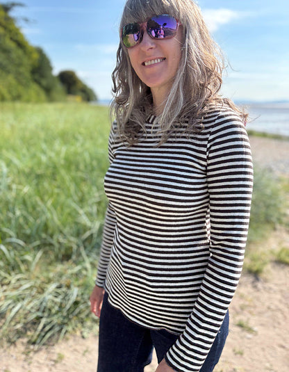 Woman wearing a black and white striped long-sleeve shirt standing outdoors with greenery and a body of water in the background.