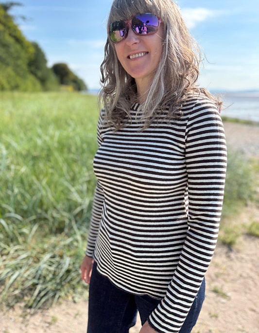 Woman wearing a black and white striped long-sleeve shirt standing outdoors with greenery and a body of water in the background.