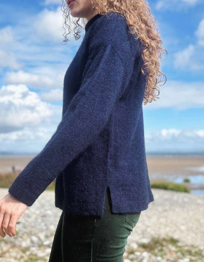 A woman wearing a dark navy blue textured knit sweater and dark green pants, standing outdoors on a pebble beach with a blue sky and clouds in the background.
