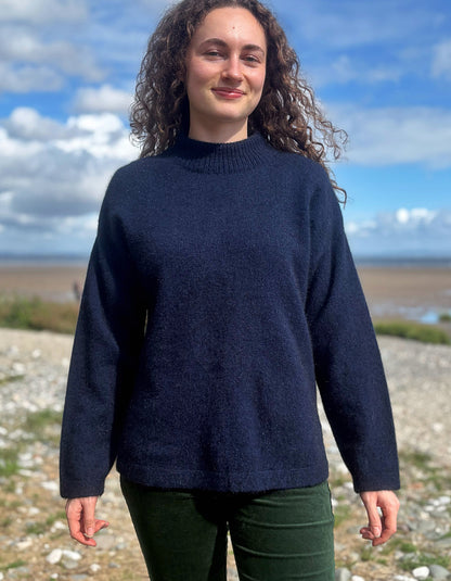 A woman with curly brown hair wearing a dark navy blue knit sweater and dark green pants, standing outdoors on a rocky shoreline with a cloudy blue sky in the background.