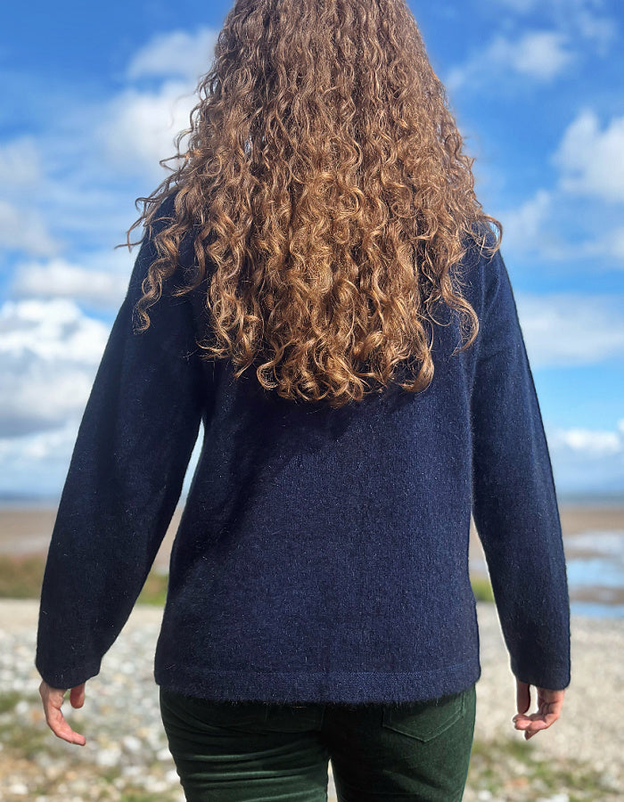 A person viewed from behind wearing a dark navy blue long-sleeve knit sweater and dark green pants, standing outdoors on a rocky shoreline under a blue sky with clouds.