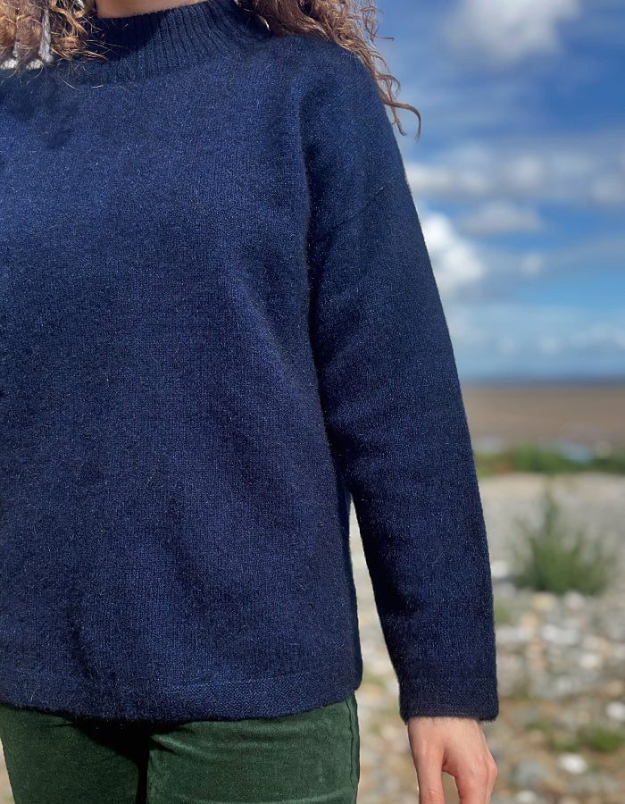 A person wearing a dark navy blue knit sweater outdoors, with a blurred natural background of blue sky, clouds, and rocky ground.