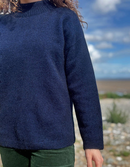 A person wearing a dark navy blue knit sweater outdoors, with a blurred natural background of blue sky, clouds, and rocky ground.