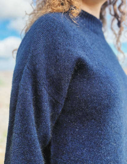 Close-up of a person wearing a textured navy blue knit sweater, showing the shoulder and upper chest against a softly blurred outdoor background.