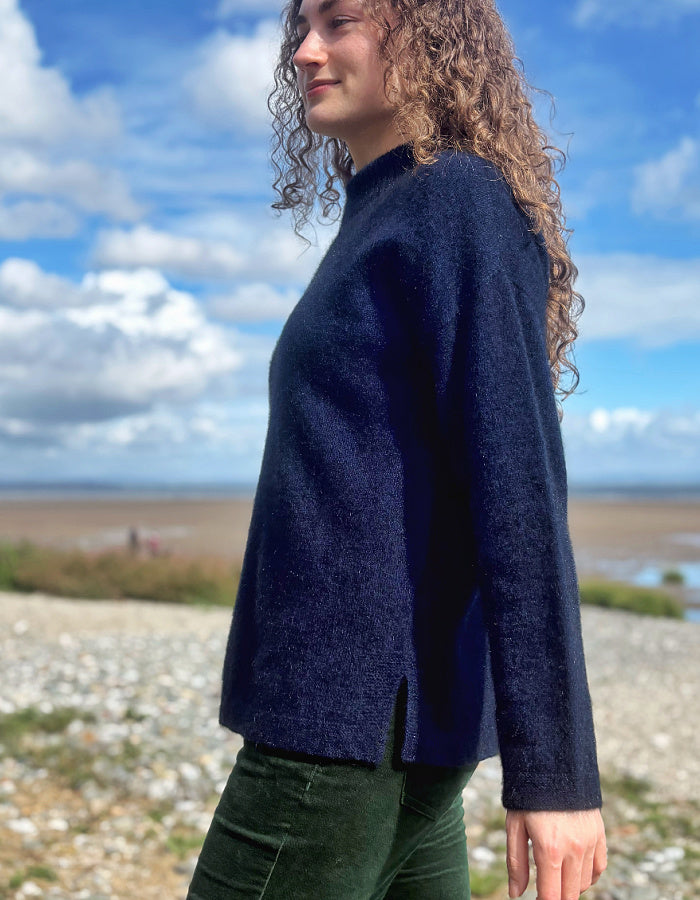 A woman with curly brown hair wearing a dark navy blue knit sweater and green pants, standing outdoors on a rocky shoreline under a blue sky with clouds.