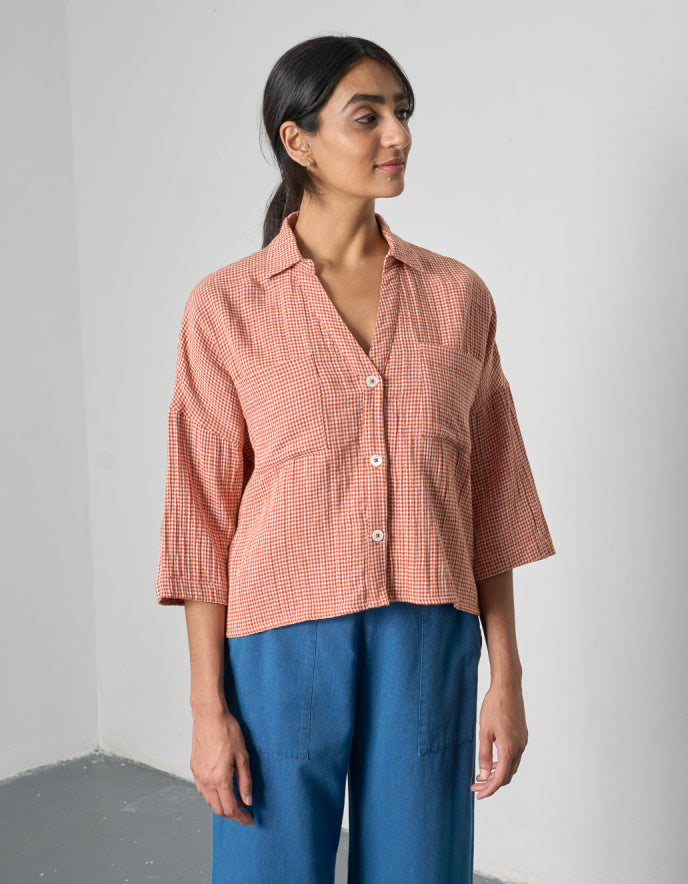 Woman wearing a pink checkered outfit standing outdoors with a wooden fence and trees in the 