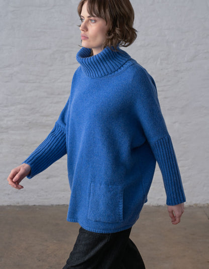 A woman wearing a vibrant blue turtleneck sweater with ribbed sleeves and a front patch pocket, standing against a white brick wall.