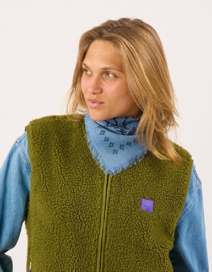 A woman wearing an olive green sherpa fleece zip-up vest over a light blue denim shirt and a blue patterned bandana, photographed against a plain white studio background.