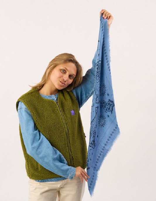 A woman modeling a sage green sherpa fleece vest over a light blue long-sleeve shirt, holding up a light blue patterned scarf with fringe.