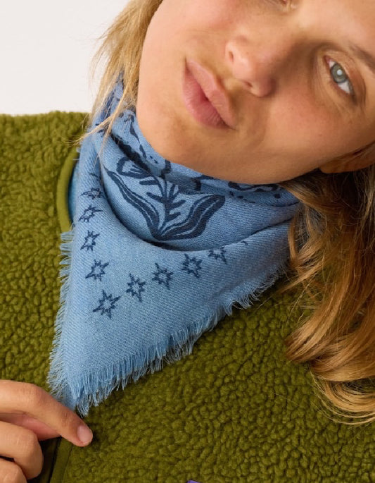 A close-up lifestyle shot of a person wearing a light blue knit bandana scarf with dark blue floral and star patterns, styled over an olive green fleece jacket.