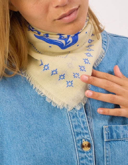 A close-up of a person wearing a cream-colored fringed bandana with blue patterns, tucked into a light blue denim shirt.