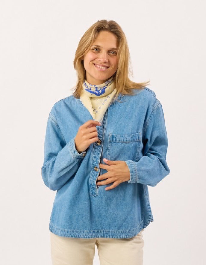 A woman wearing a light blue denim button-up shirt over a cream turtleneck with a blue patterned scarf, paired with beige pants, photographed against a plain white studio background.