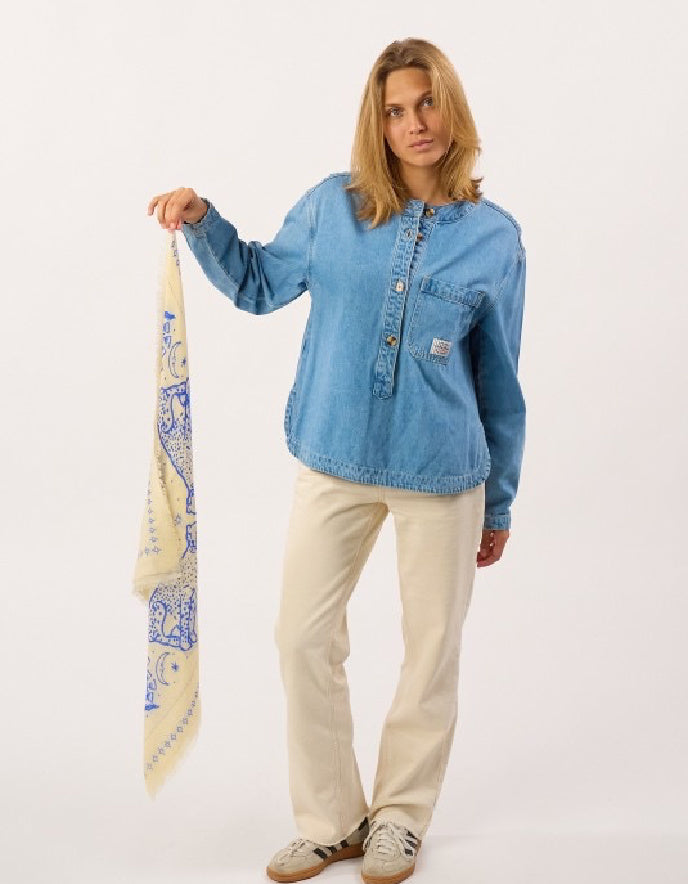 A woman modeling a light blue denim shirt-jacket with a small Carhartt brand patch on the chest pocket, paired with cream pants and holding a cream scarf with a blue paisley pattern against a plain white studio background.