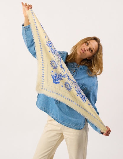 A woman wearing a light blue denim shirt and cream pants holds up a large cream-colored square scarf with blue patterns against a plain white background.