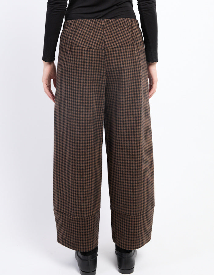 A person shown from the back wearing brown and black checkered wide-leg cropped trousers, a black long-sleeve top, and black shoes against a plain white studio background.