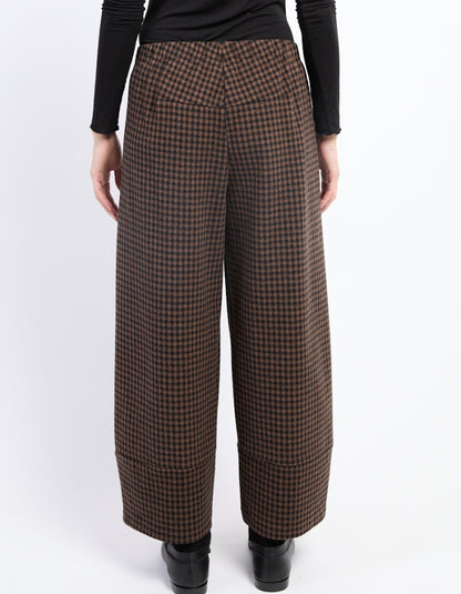 A person shown from the back wearing brown and black checkered wide-leg cropped trousers, a black long-sleeve top, and black shoes against a plain white studio background.