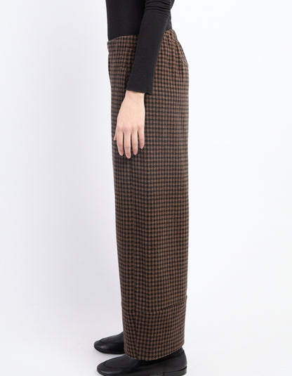 A side view of a person wearing brown and black checkered wide-leg trousers, a black long-sleeve top, and black shoes against a plain white background.