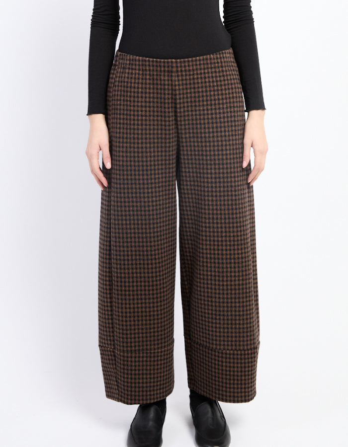 A model wearing brown and black checkered wide-leg cropped trousers, paired with a black long-sleeve top and black boots, against a plain white studio background.