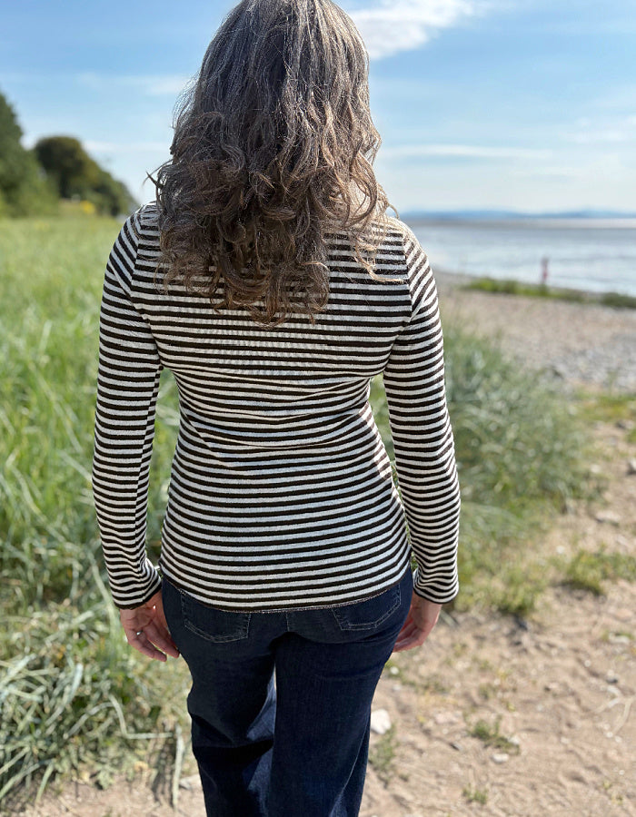 Woman wearing a black and white striped long-sleeve shirt standing outdoors with greenery and a body of water in the background.