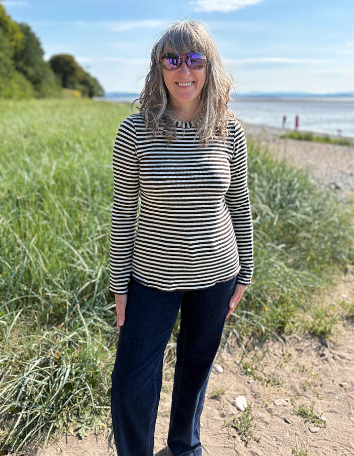Woman wearing a black and white striped long-sleeve shirt standing outdoors with greenery and a body of water in the background.