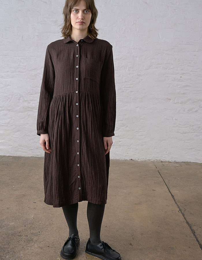 A woman wearing a dark brown crinkle-textured button-down shirt dress, black tights, and black leather shoes, standing against a white brick wall.