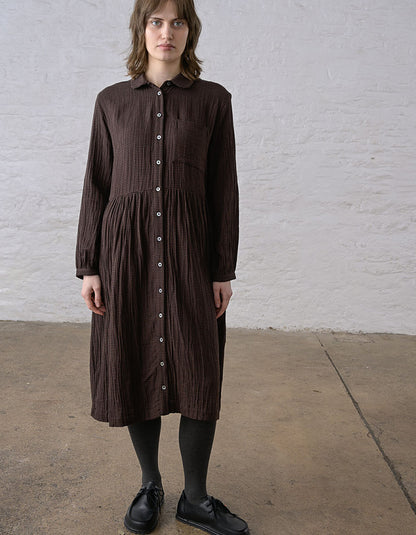 A woman wearing a dark brown crinkle-textured button-down shirt dress, black tights, and black leather shoes, standing against a white brick wall.