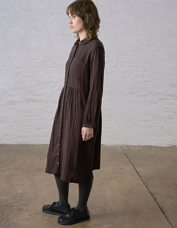 A woman wearing a dark brown crinkled midi shirt dress with long sleeves and a button-down front, standing in profile against a white brick wall.