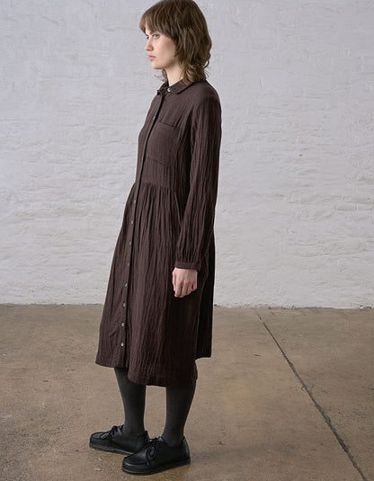 A woman wearing a dark brown crinkled midi shirt dress with long sleeves and a button-down front, standing in profile against a white brick wall.