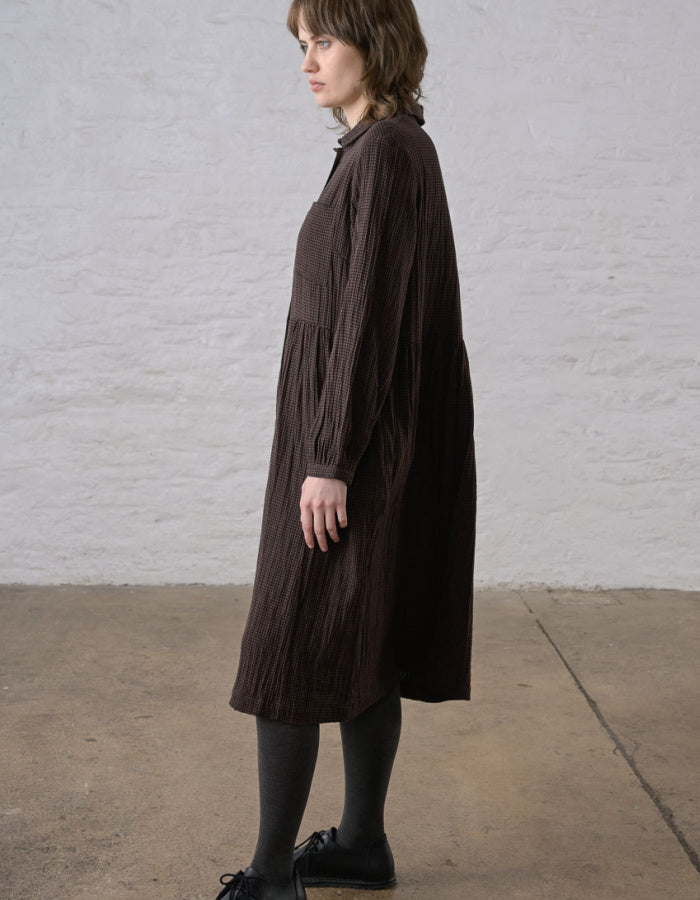 A woman wearing a dark brown crinkled midi dress, standing in profile against a white brick wall and concrete floor.