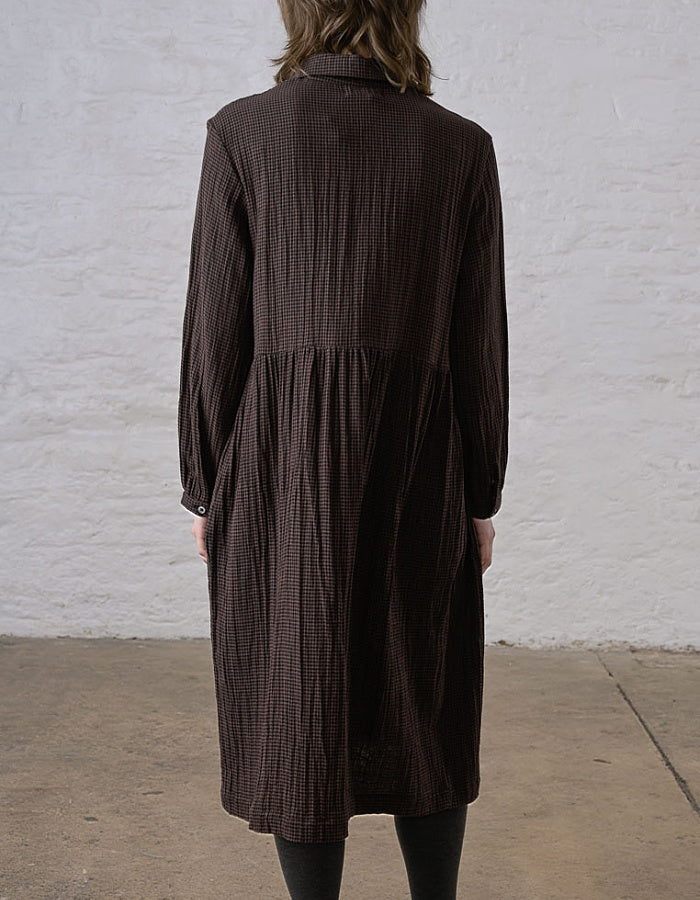 A person viewed from the back wearing a dark brown long-sleeve midi dress with a subtle textured grid pattern, standing against a white brick wall.