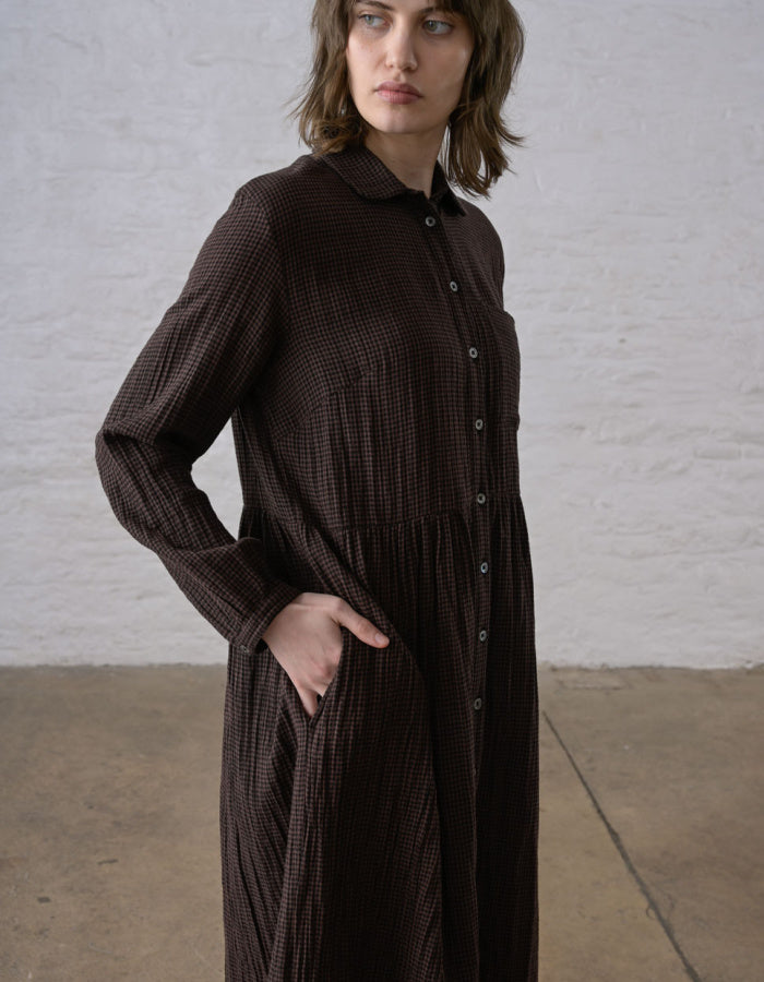 A woman wearing a dark brown textured long-sleeve button-up dress, standing against a white brick wall.
