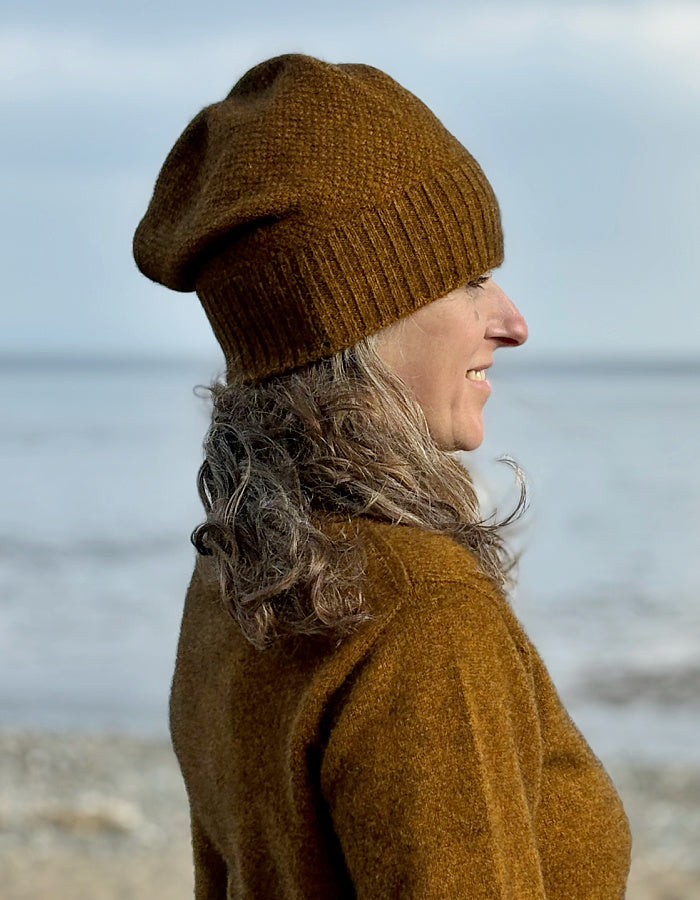 A woman in profile wearing a brown knit slouchy beanie and a matching brown sweater, standing outdoors with a blurred beach or water background.