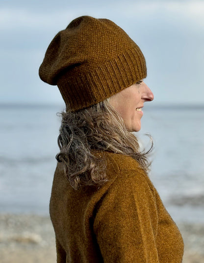 A woman in profile wearing a brown knit slouchy beanie and a matching brown sweater, standing outdoors with a blurred beach or water background.