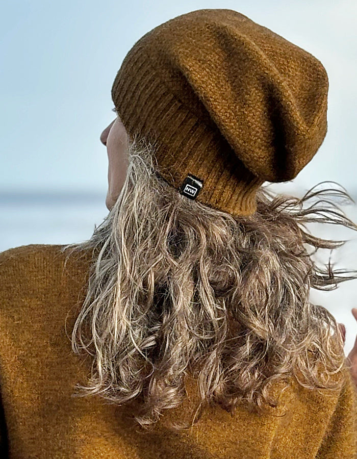 A person shown from the back wearing a textured mustard-brown knit beanie and a matching brown sweater, with wavy light brown hair blowing in the wind against a pale sky background.